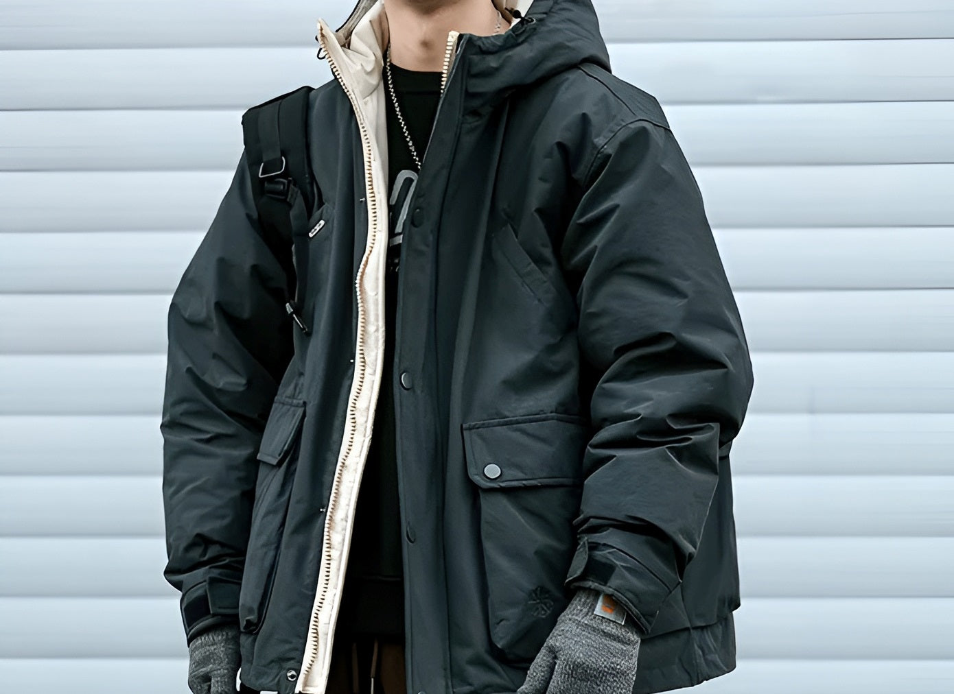 Man wearing a black hooded jacket layered over a beige inner zip-up and black shirt, styled with gray gloves and a backpack.