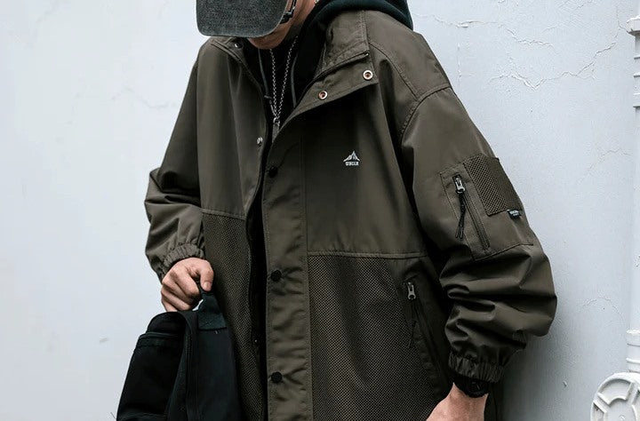 Person wearing an olive green hooded jacket with mesh details and a cap, holding a black bag.