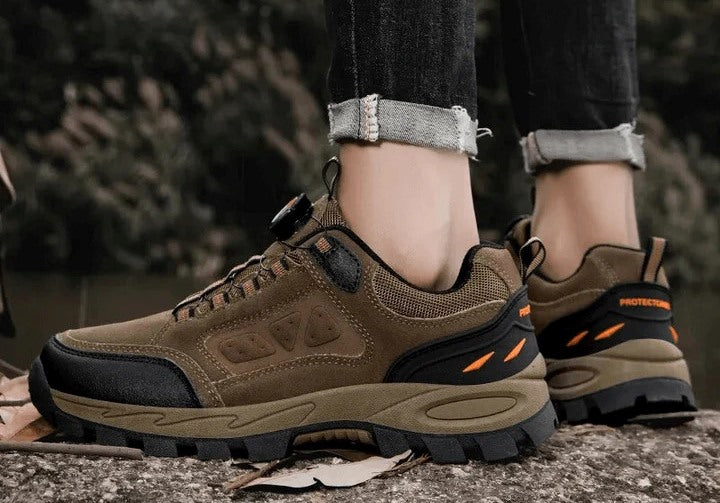 Close-up of a pair of rugged brown hiking shoes with orange accents, worn by a person standing on a trail, showcasing the shoes' durable sole and design.