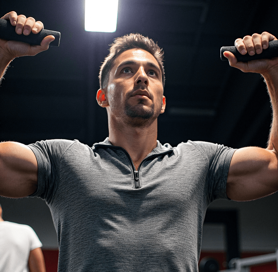 A man wearing a gray zip-up athletic T-shirt, lifting weights in a gym while focusing intently. The lighting highlights his muscular build.