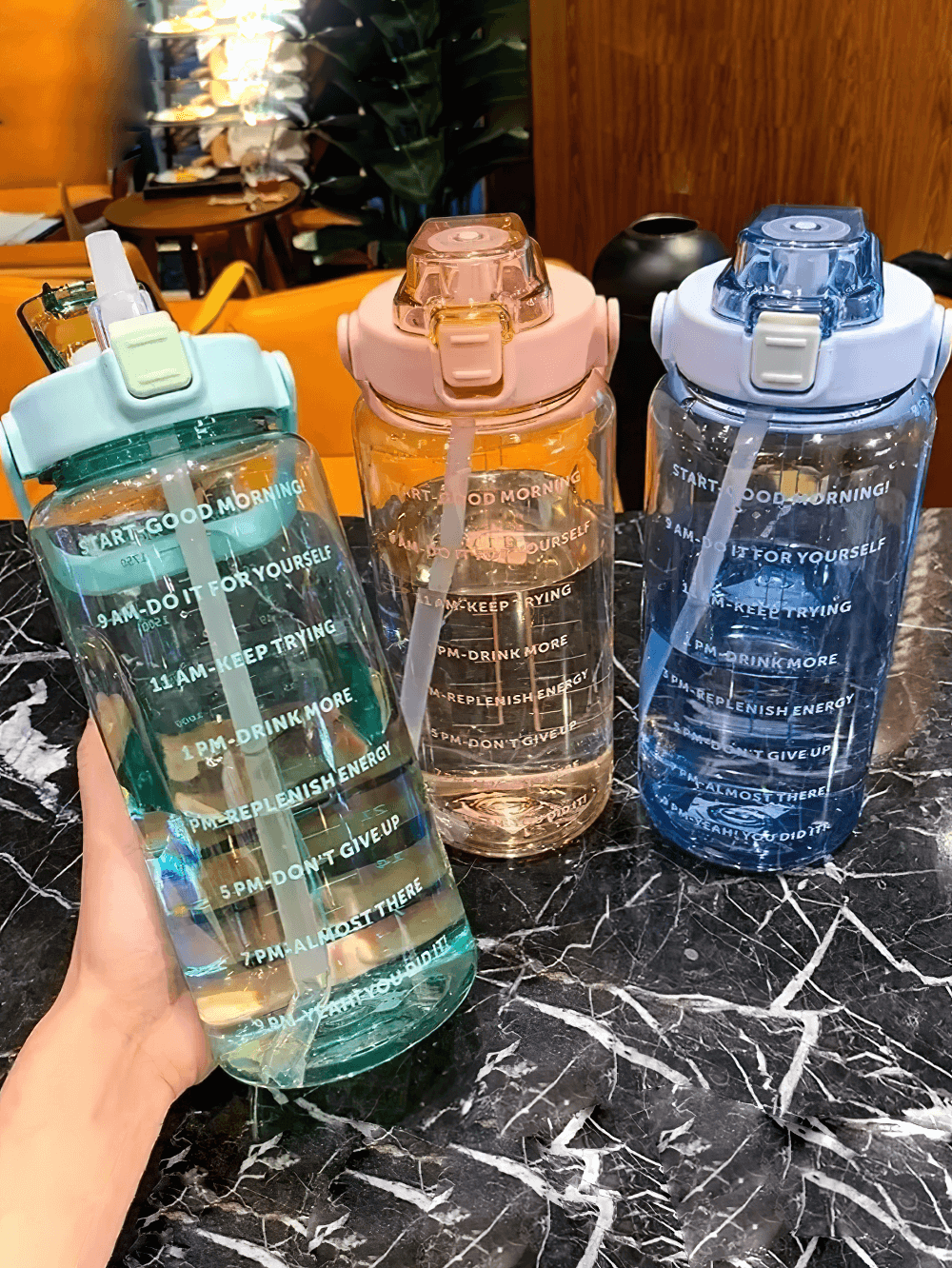 Three stylish 2L sports water bottles with straws in blue, pink, and aqua on a marble table, featuring motivational time markers.