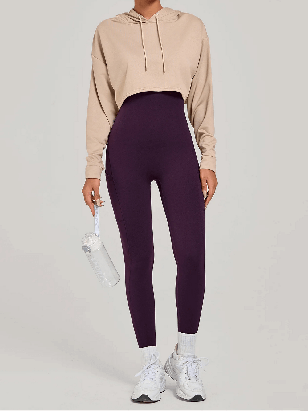 Woman in beige hoodie and purple leggings holding water bottle, showcasing stylish activewear look.