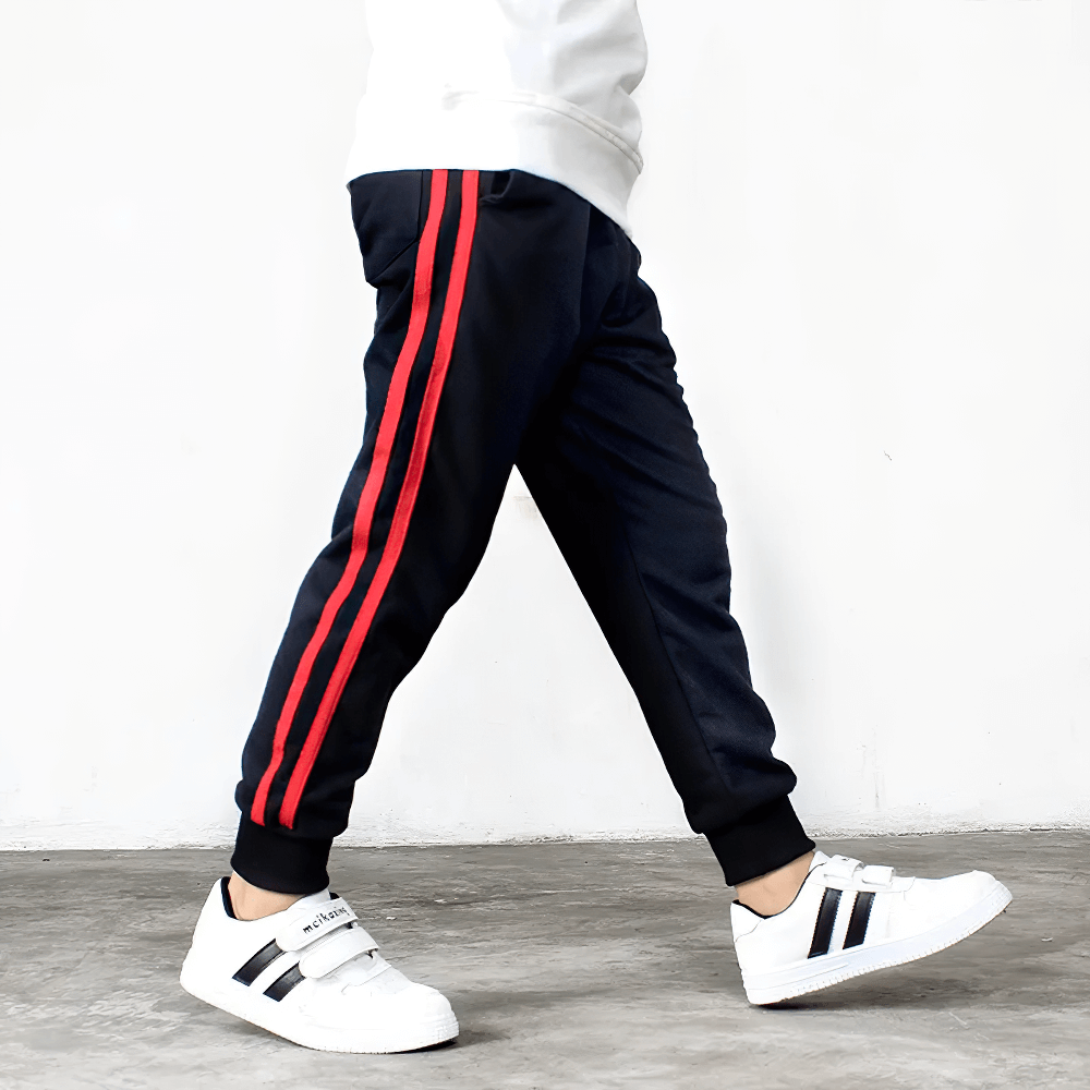 Boy wearing Boys Athletic Jogger Sweatpants with Pockets - SF3611, featuring red side stripes and white sneakers, walking on concrete floor.