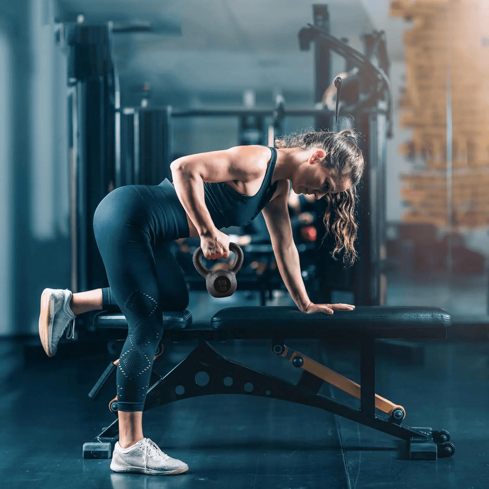 Woman using kettlebell for strength training workout in gym, demonstrating proper form and balance on bench.