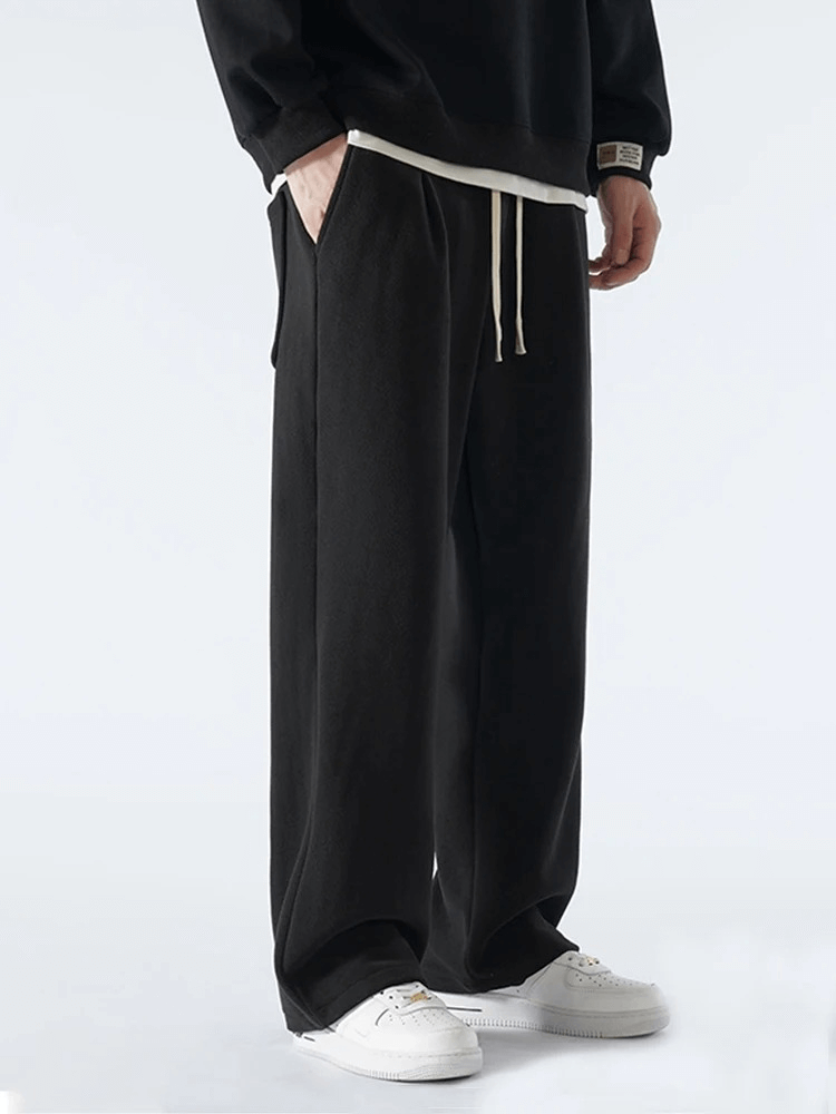 Men's casual wide fleece sweatpants with mid-rise waist, drawstring, side pockets, and loose fit in black, paired with white sneakers.