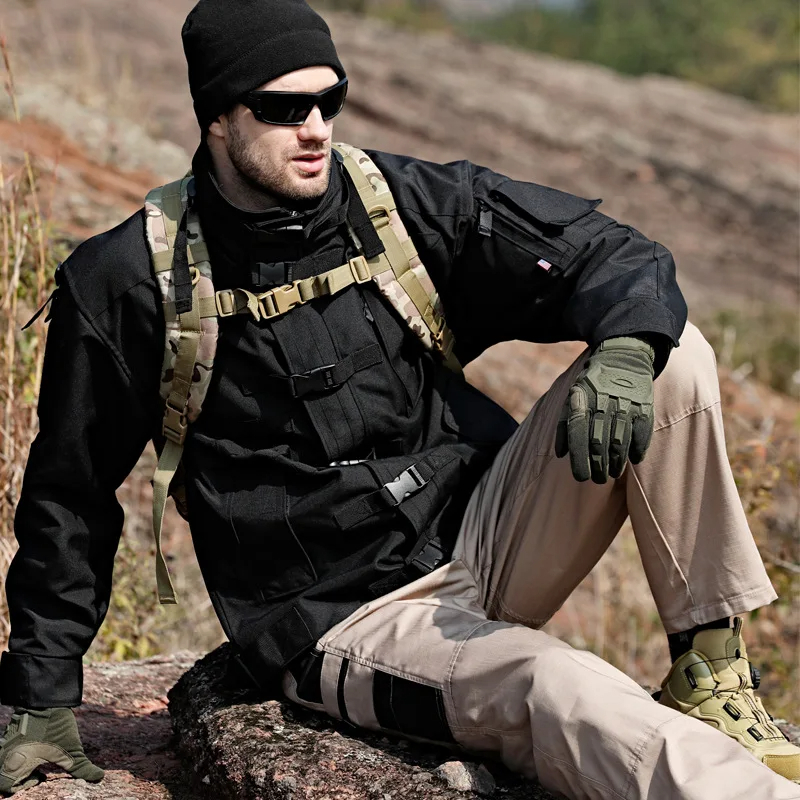 Man wearing tactical cargo pants and jacket, equipped for hiking, resting on a rock in outdoor rugged terrain.