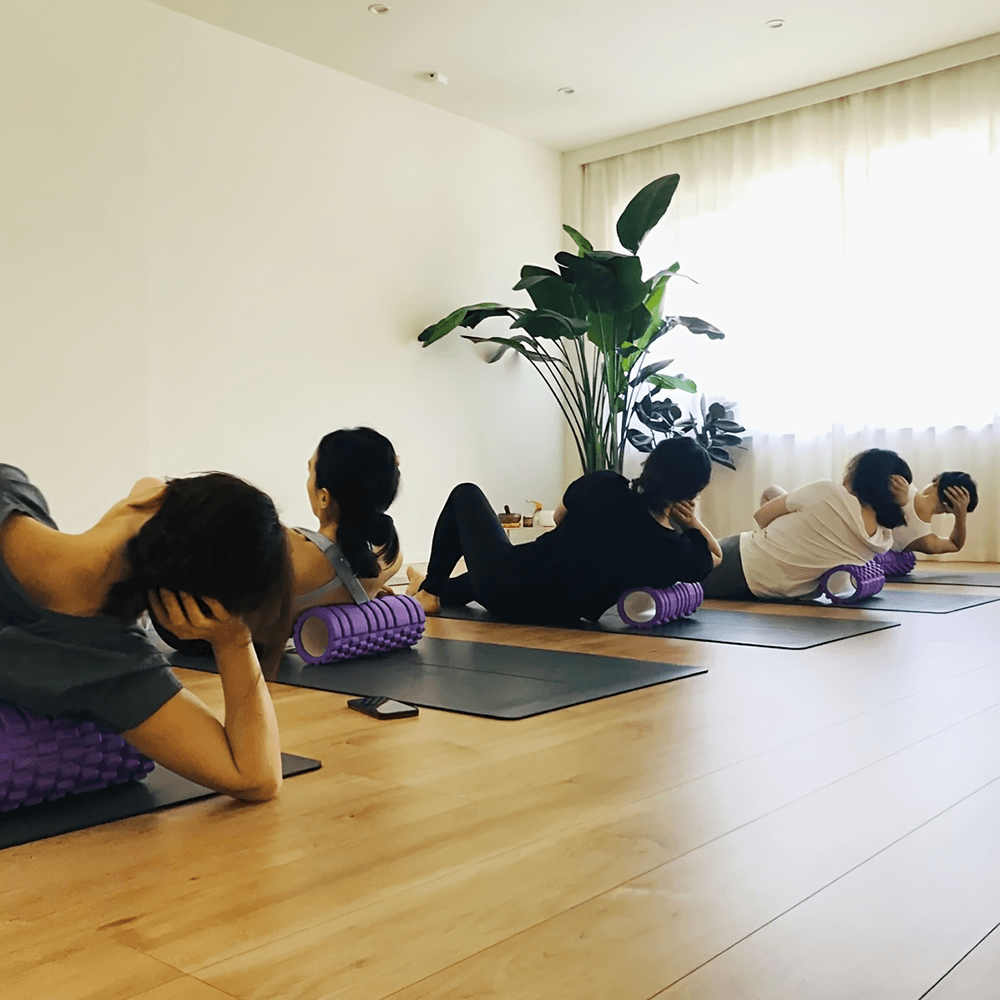 Group yoga class using EVA foam rollers for deep tissue massage and myofascial release exercises on mats in a sunlit room.