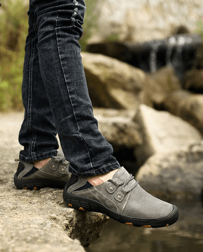 Fashionable breathable leather trekking shoes SF1344 on man standing on rocky terrain near waterfall, showcasing comfort and durability.