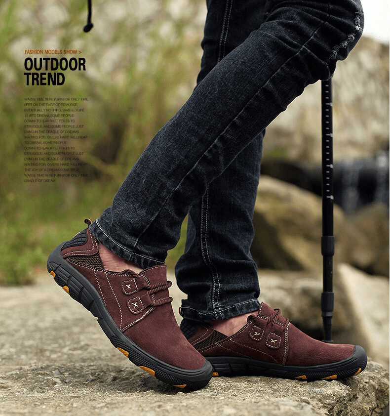 Man wearing brown leather trekking shoes with rubber outsole for outdoor adventure, standing on rocky path with trekking pole.