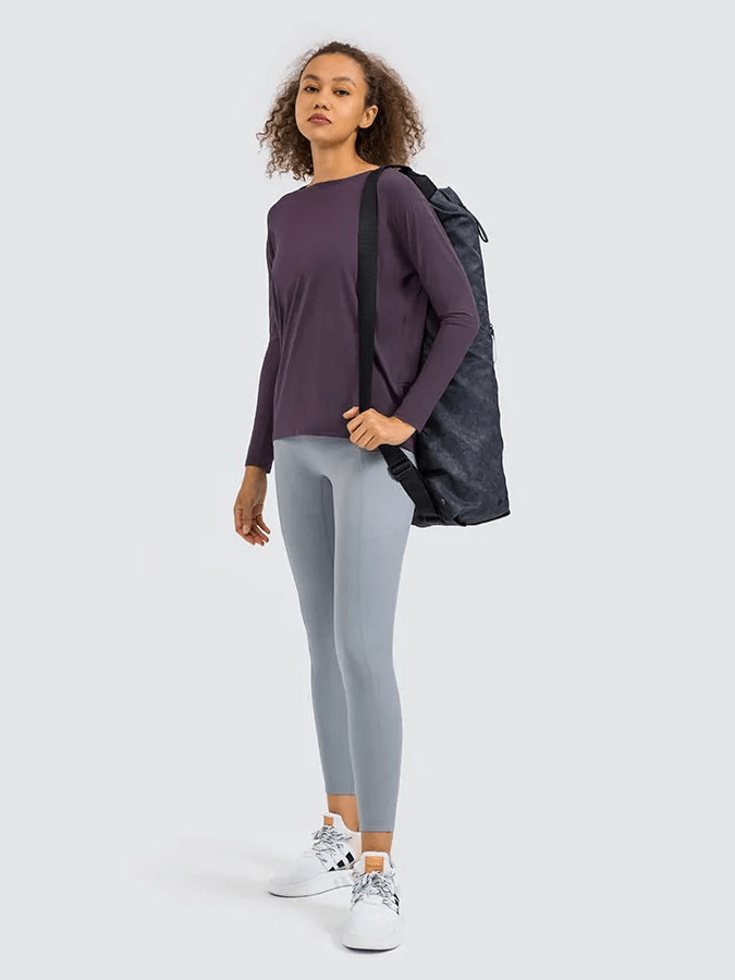 Sporty woman wearing a solid color long sleeves loose top for gym with leggings, carrying a gym bag, isolated on gray background.