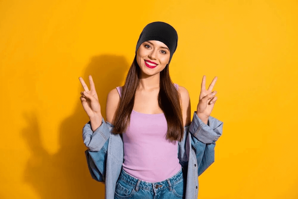 Smiling person in black beanie posing against yellow background, wearing casual denim and pink top, displaying peace signs.