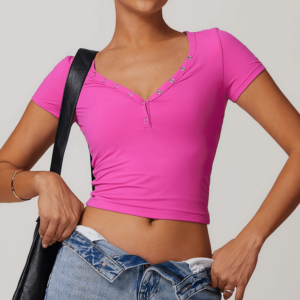 Woman wearing a pink V-neck button short sleeve yoga T-shirt with a fitted design, holding jeans and a black bag, showcasing a casual look.