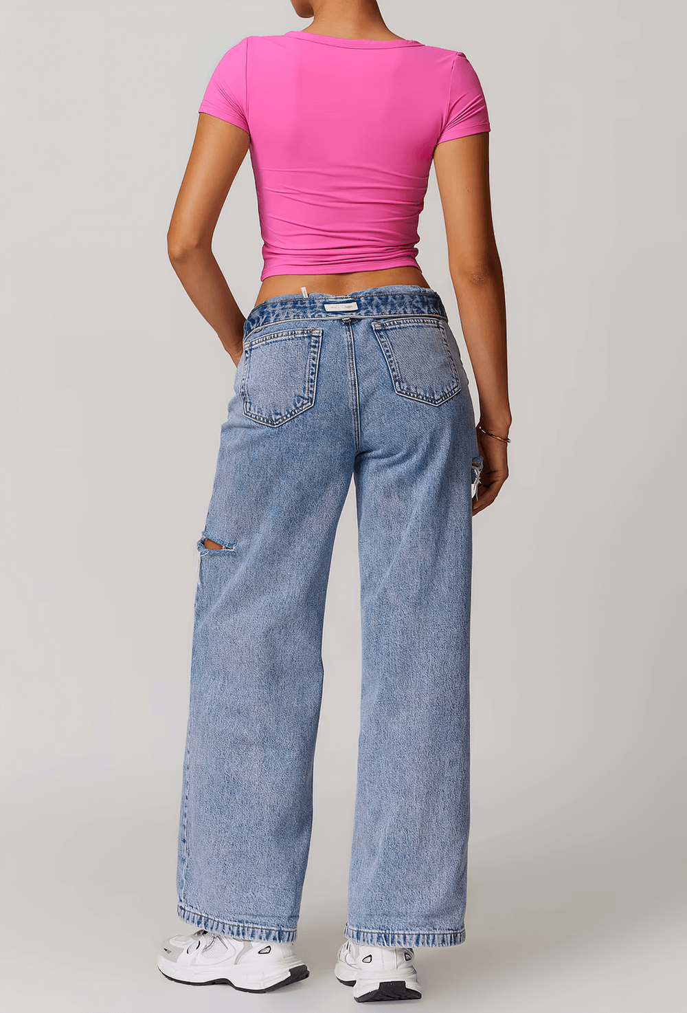 Woman in pink crop top and blue jeans, standing with back to camera, showcasing casual style with athletic white sneakers.