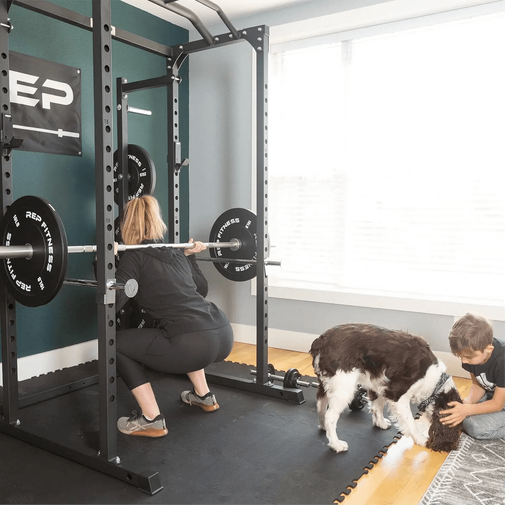 Woman lifting Barbell with REP Fitness Bumper Plates in home gym, child and dog nearby. Strength training equipment for workouts.