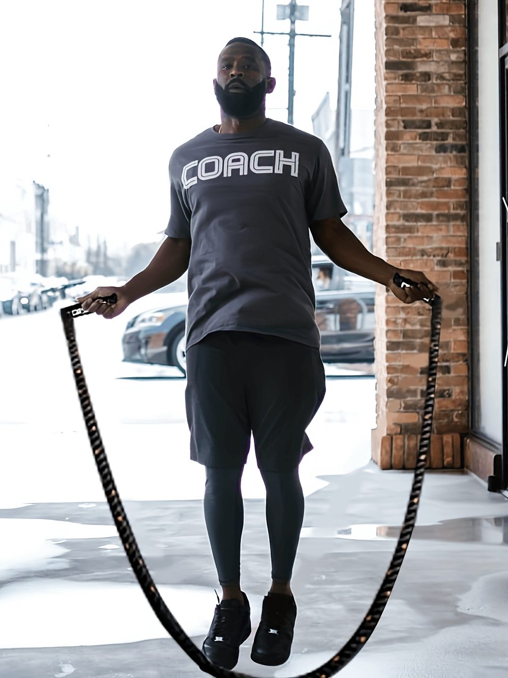 Man jumping rope with black weighted battle rope in urban setting, wearing COACH shirt, focusing on strength and endurance training.