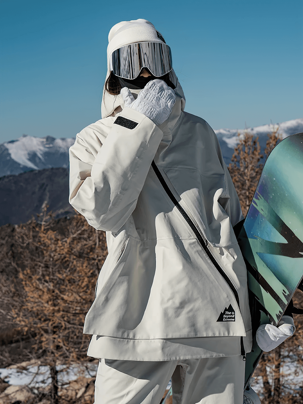 Woman wearing white hooded jacket with asymmetric zipper, posing with snowboard in snowy mountains; stylish and functional winter wear.