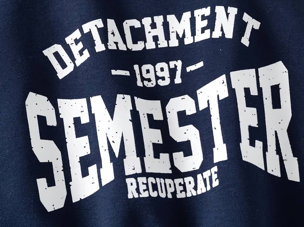 Close-up of 'Detachment Semester 1997 Recuperate' print on kid's casual sweatshirt, featuring bold white lettering on navy fabric.