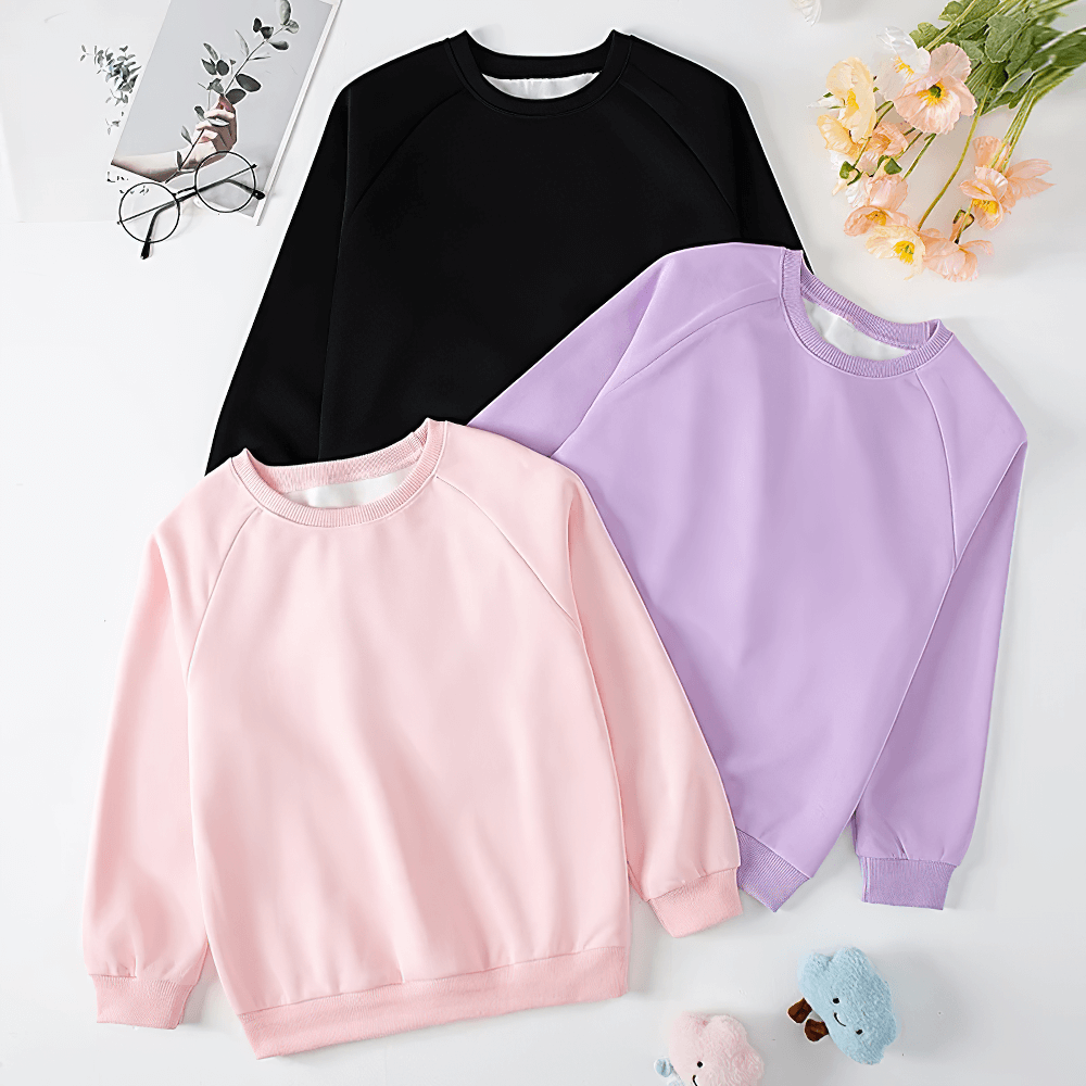 Three kids' solid color sweatshirts in black, pink, and purple, laid flat on a white background with flowers and toys.