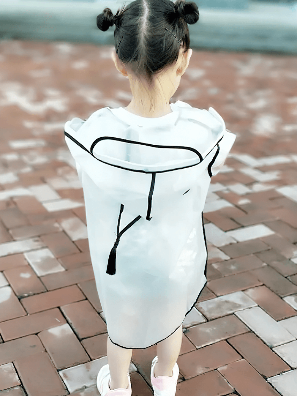 Child wearing Kids Transparent Long Hooded Raincoat - SF3671 with hood and black trim, standing on a brick path.