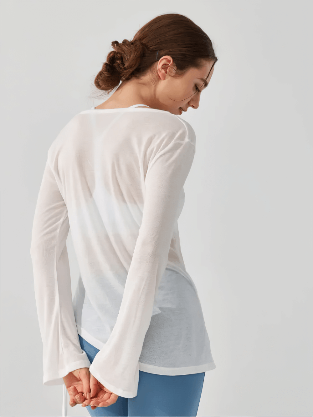 Woman in back view wearing white long-sleeve yoga top with ruched side-tie detail over blue leggings.