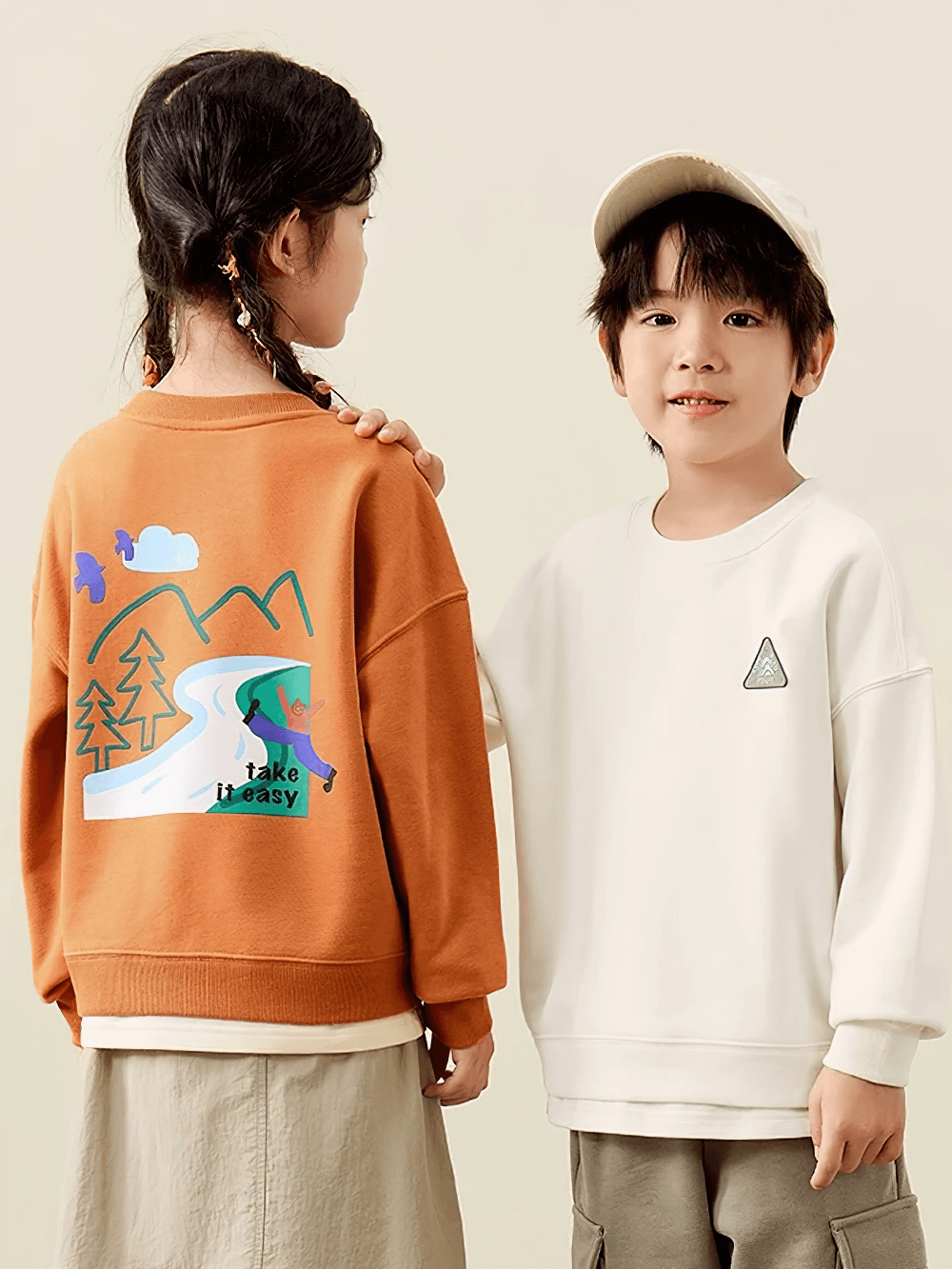 Two kids wearing trendy long-sleeved sweatshirts; one with a mountain design and 'Take it Easy' text, perfect for adventurous spirits.