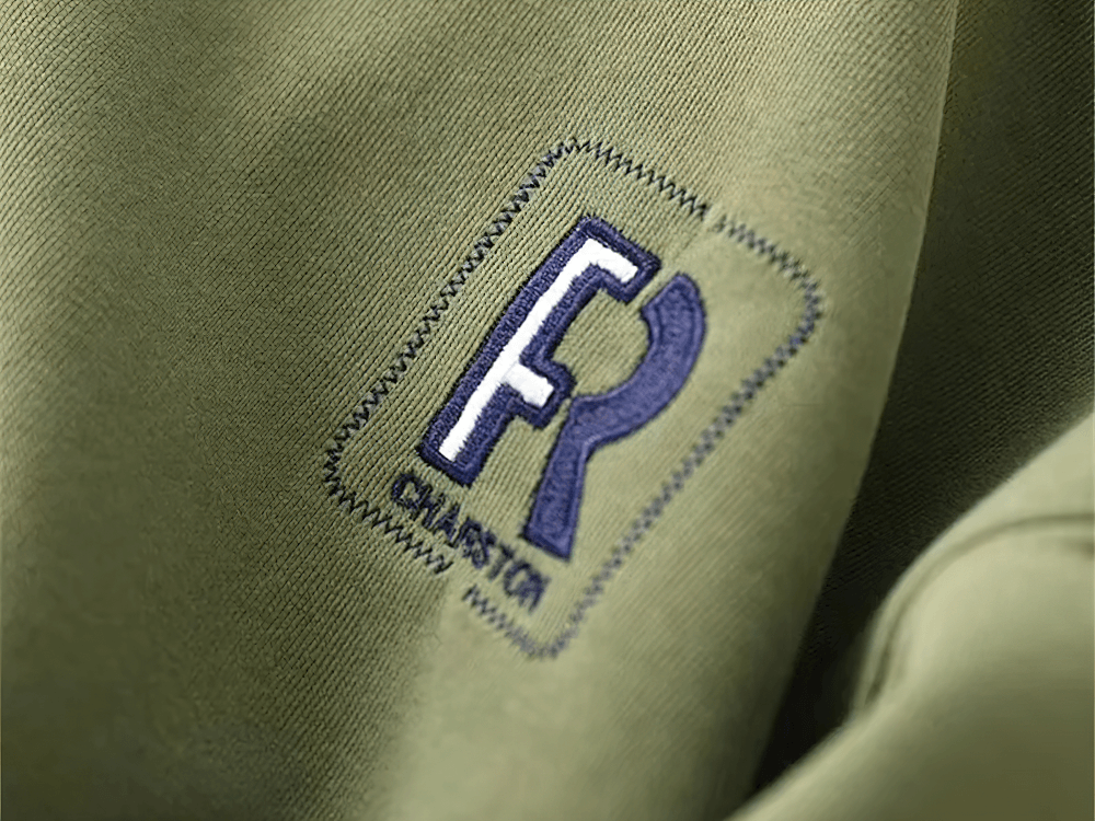Close-up of an olive green fabric with an embroidered logo, depicting the letters 'FR' and 'CHASRTA' in blue and black.