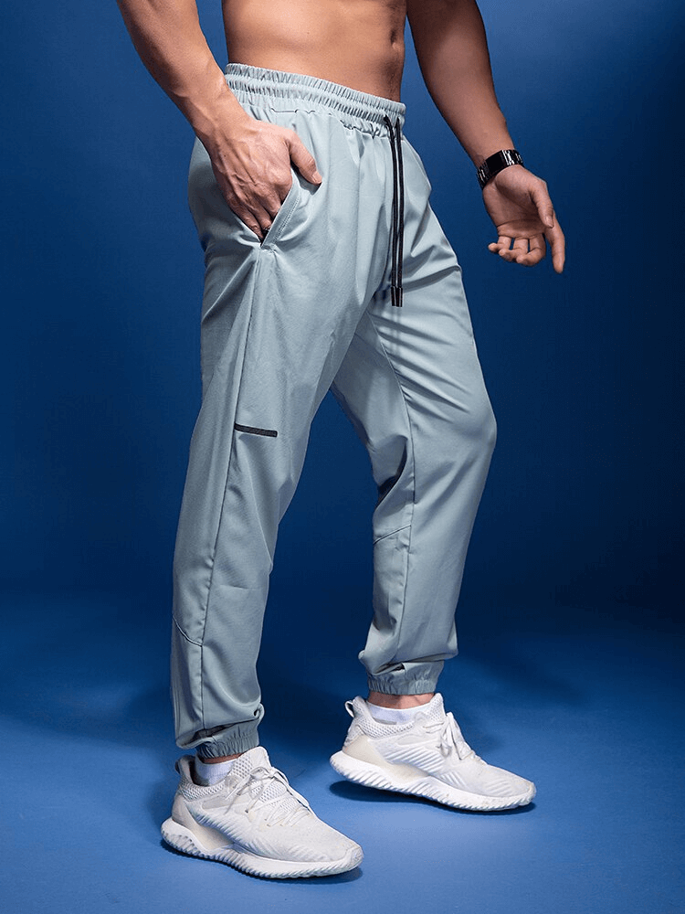 Male running joggers with elastic waist and zipper pockets, blue backdrop, ideal for sports and active lifestyle, model wearing white sneakers.