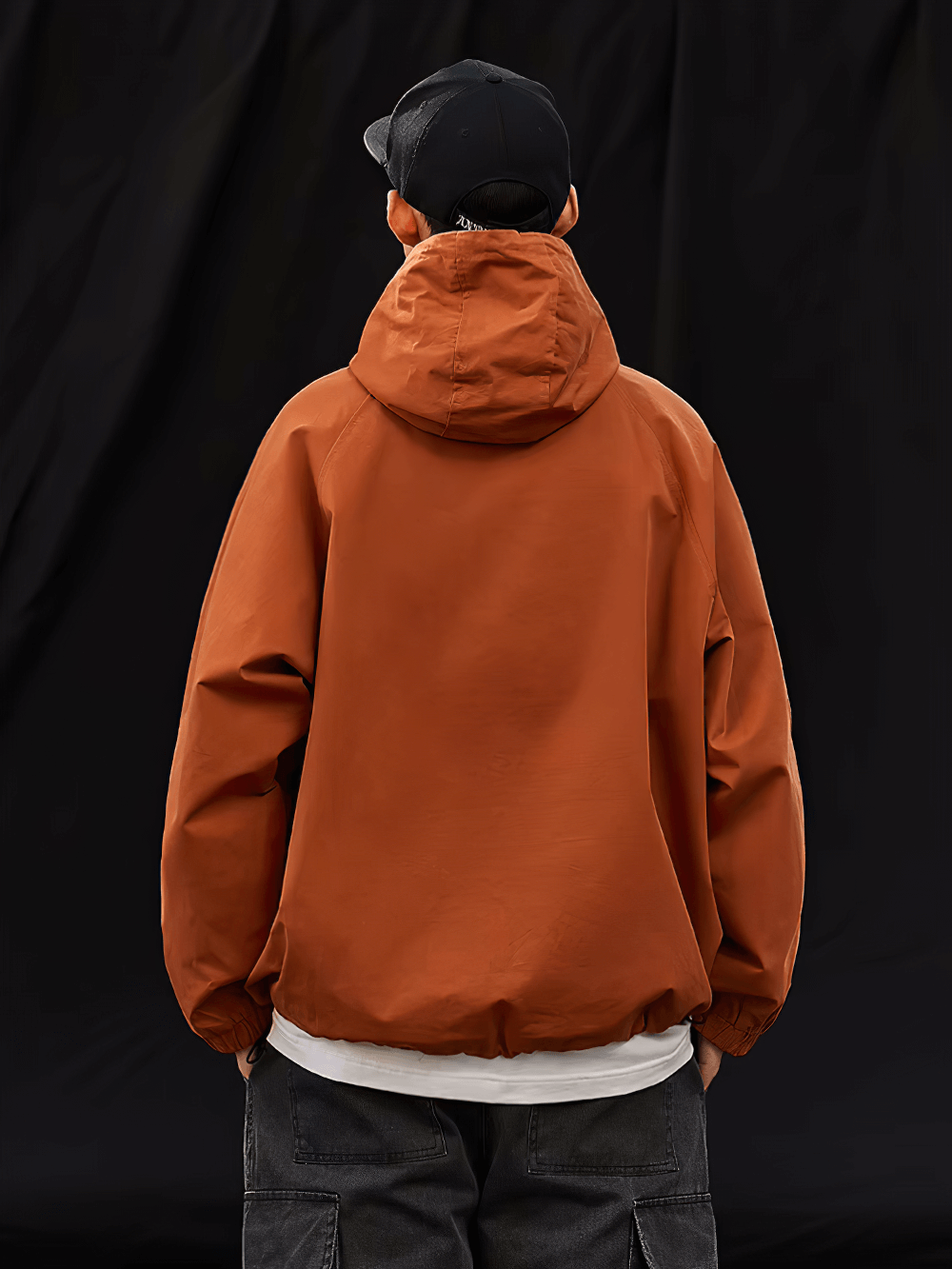 Back view of a man wearing an orange hooded pullover jacket with black cap, showcasing streetwear style.