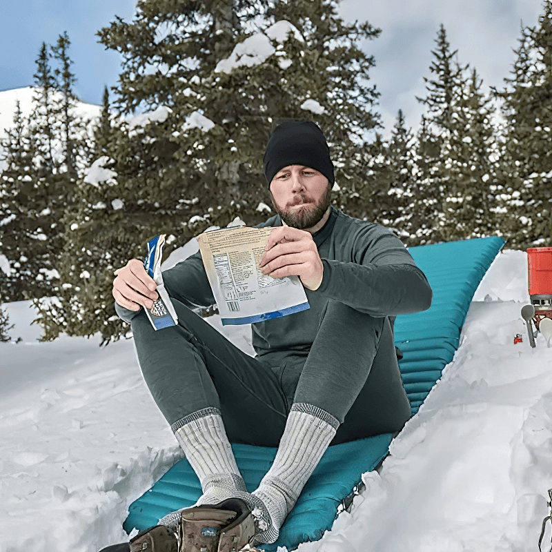 Man in merino wool thermal pants sitting on snow with a book, enjoying winter outdoors.