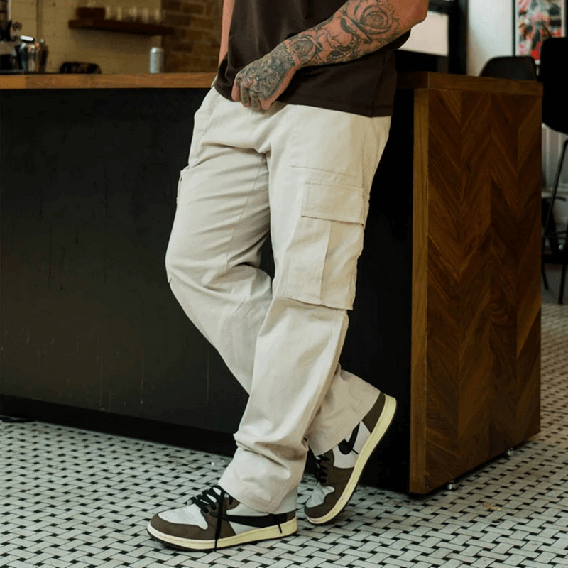 Man wearing multi-pocket training cargo trousers SF1921, beige color, leaning casually next to a wooden counter.