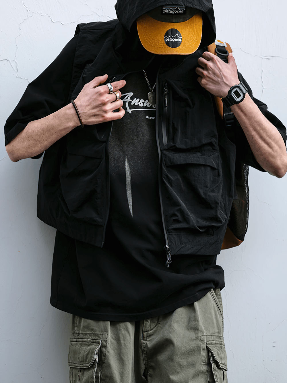 Man wearing a black multi-pocket waterproof cargo vest with hood, styled with outdoor gear.