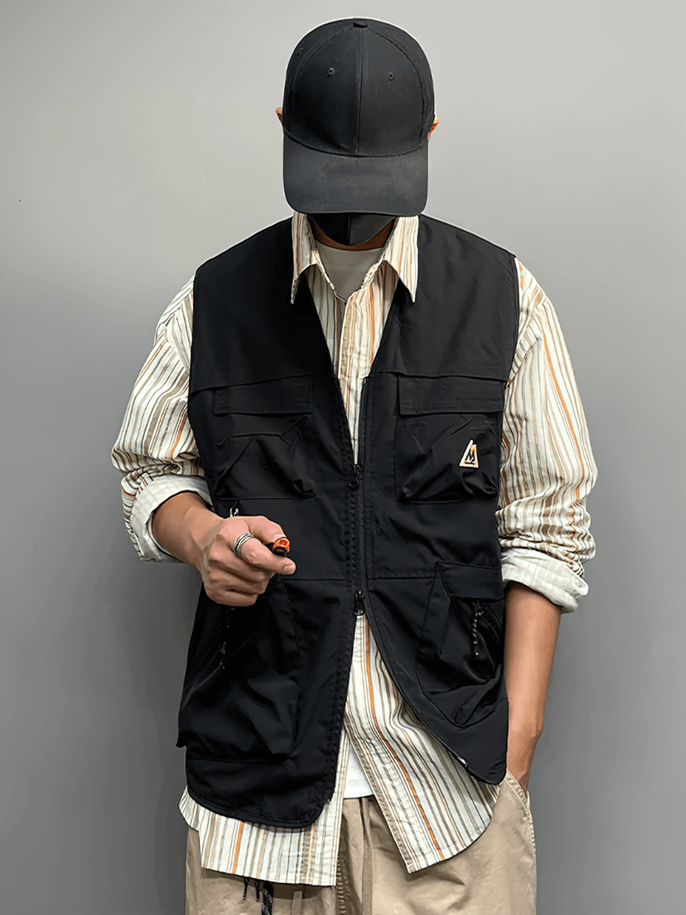 Man wearing a black tactical utility vest with multiple pockets over a striped shirt, holding a small tool, ready for adventure.