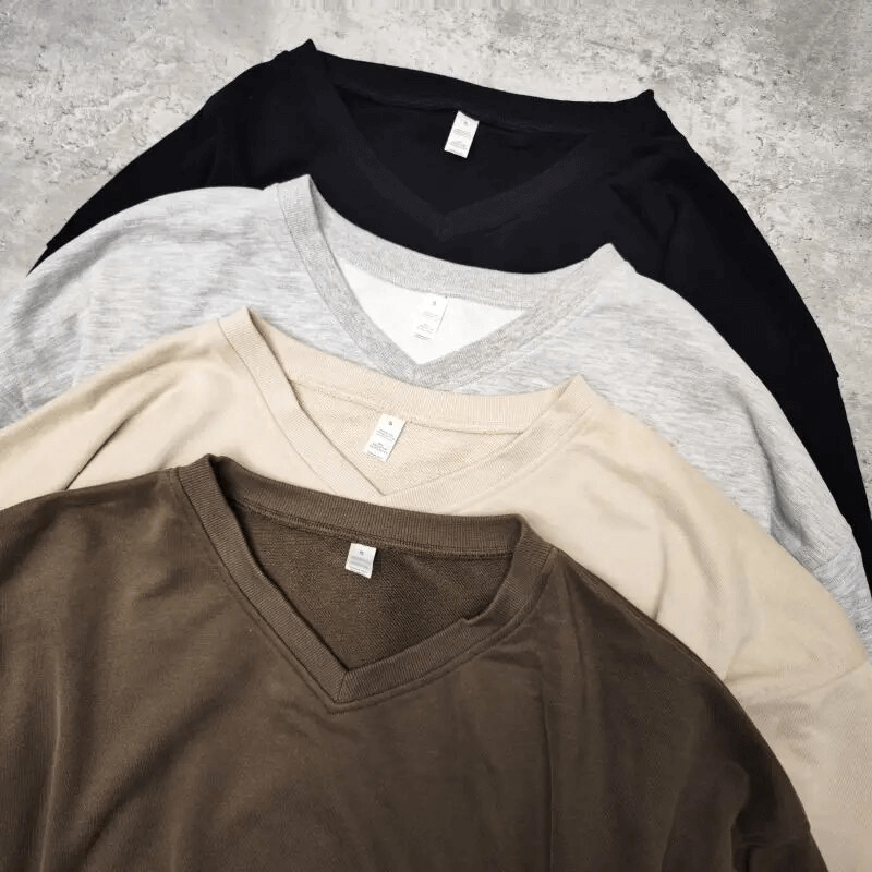 Stack of four V-neck cotton t-shirts in brown, beige, gray, and black displayed on a textured backdrop.
