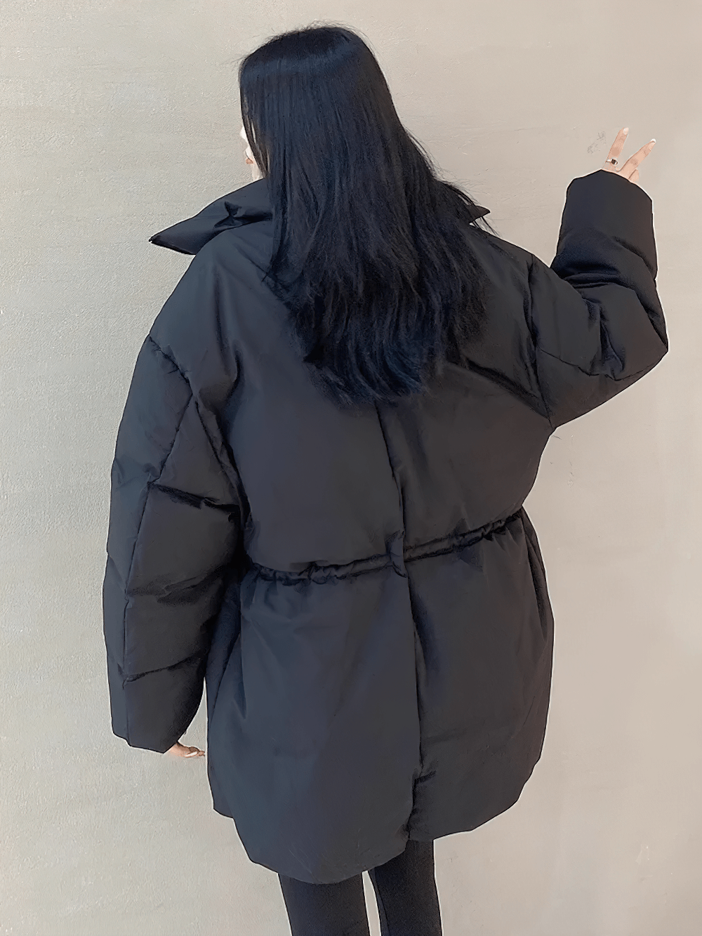 Woman wearing a stylish black puffer jacket with adjustable waist, showing a peace sign from behind against a neutral background.