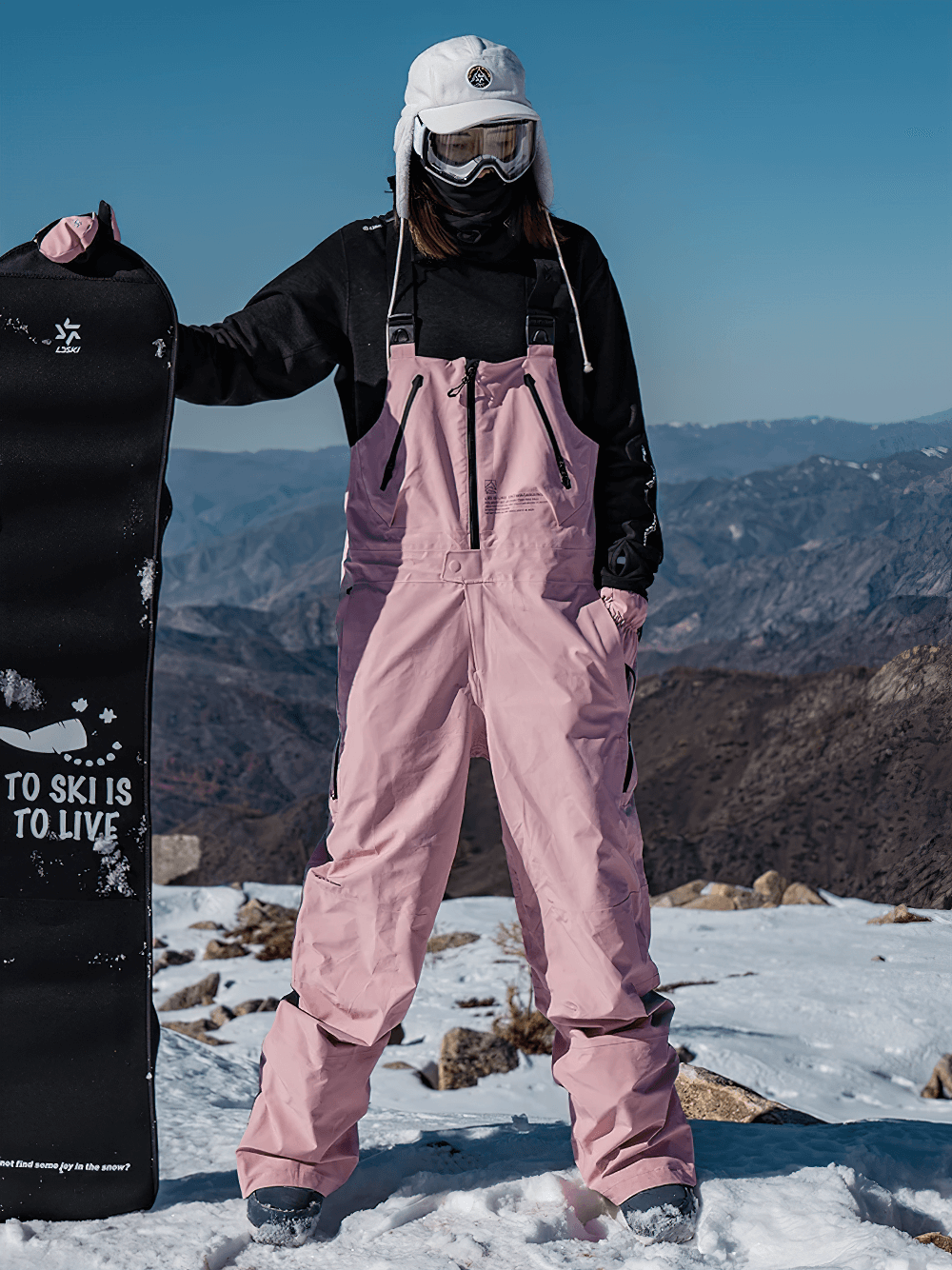 Woman wearing pink snowboard bib pants with adjustable straps, standing on snowy mountain, holding snowboard and wearing goggles.