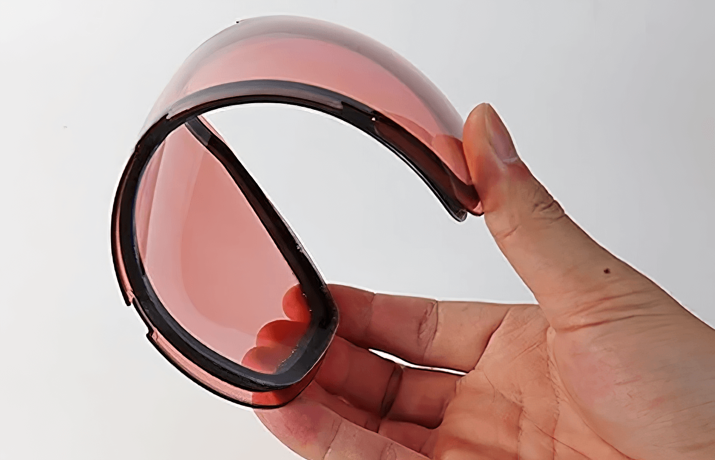 Flexible polycarbonate lens in hand, showcasing durability and light red tint for snowboarding goggles.