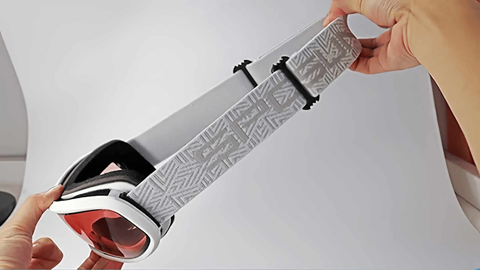 Hands holding snowboarding goggles with red-tinted lenses and patterned strap, perfect for UV protection and anti-fog skiing adventures.