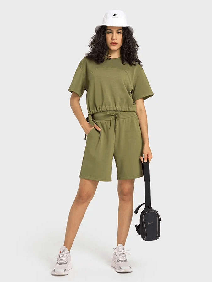 Woman wearing an olive green short-sleeve crop top and shorts with a white hat, holding a bag, on a neutral background.