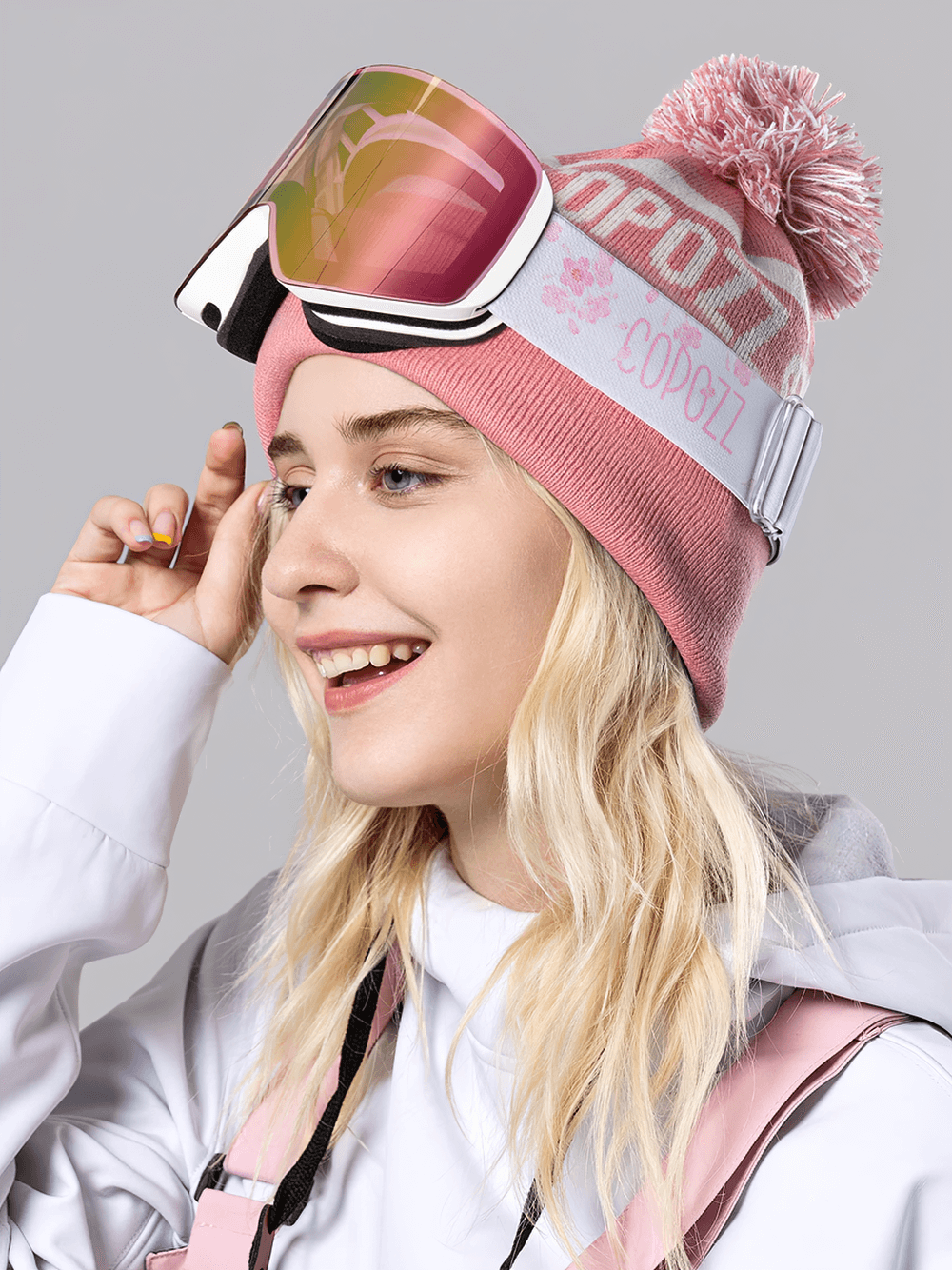 Woman wearing stylish cylindrical snowboard goggles with UV400 protection, pink beanie, and ski jacket, smiling on the slopes.