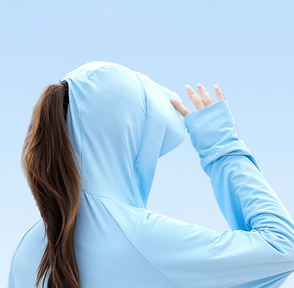 Woman wearing stylish light blue hooded UV protection sun jacket, covering face from sun with hand, against clear sky background.