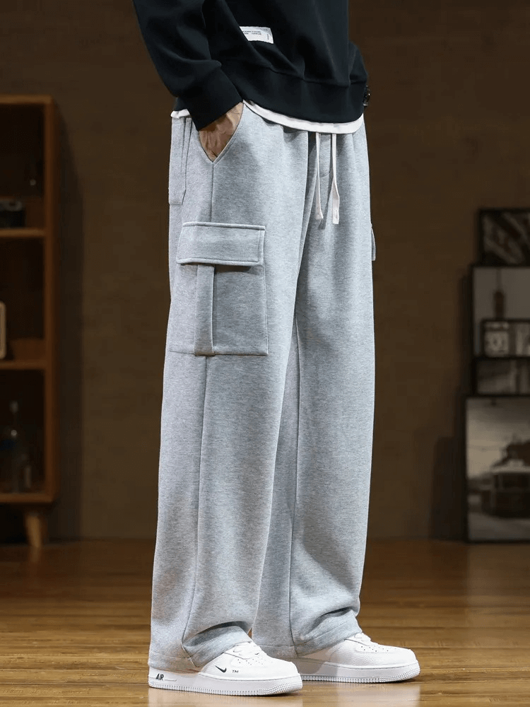 Stylish grey men's sports pants with several pockets, drawstring waist, and loose fit. Made from durable materials. Model standing indoors.