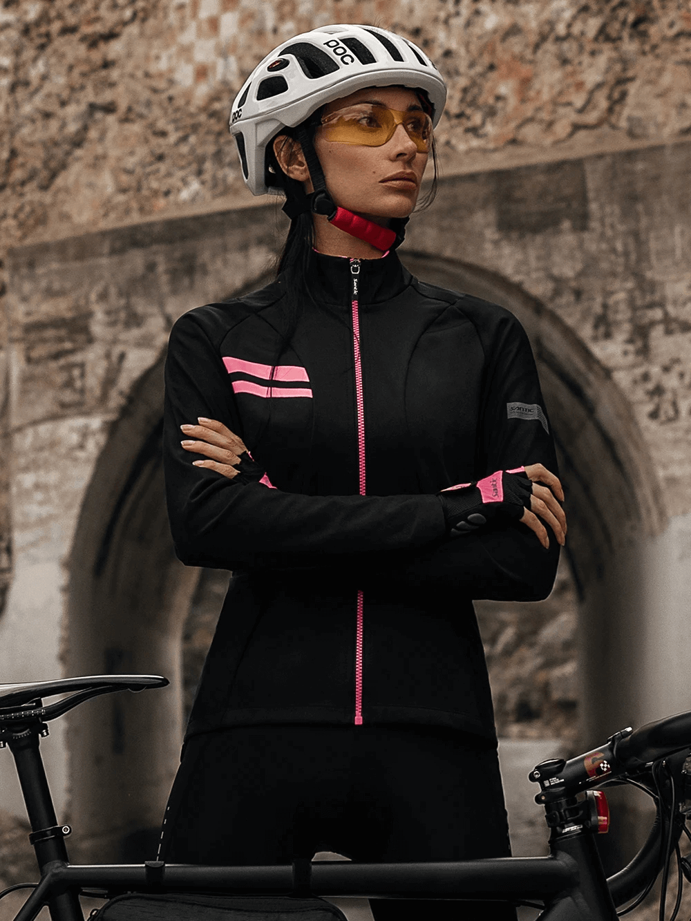 Stylish woman in a black thermal cycling jersey set with pink accents, wearing a helmet, standing confidently by her bike.