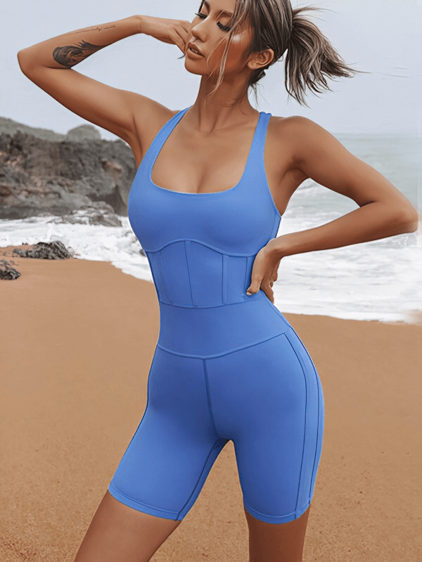 Woman wearing tight elastic one-piece workout jumpsuit for yoga, in bright blue, beach background. Perfect fit, breathable acetate.