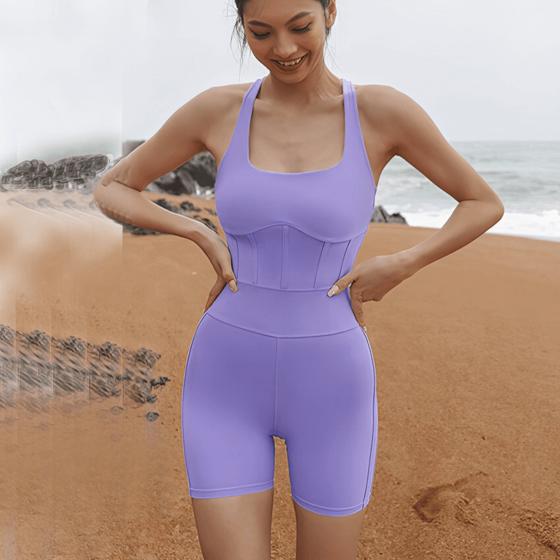 Woman wearing tight elastic one-piece workout jumpsuit, purple sleeveless design, perfect for yoga, standing on a sandy beach.