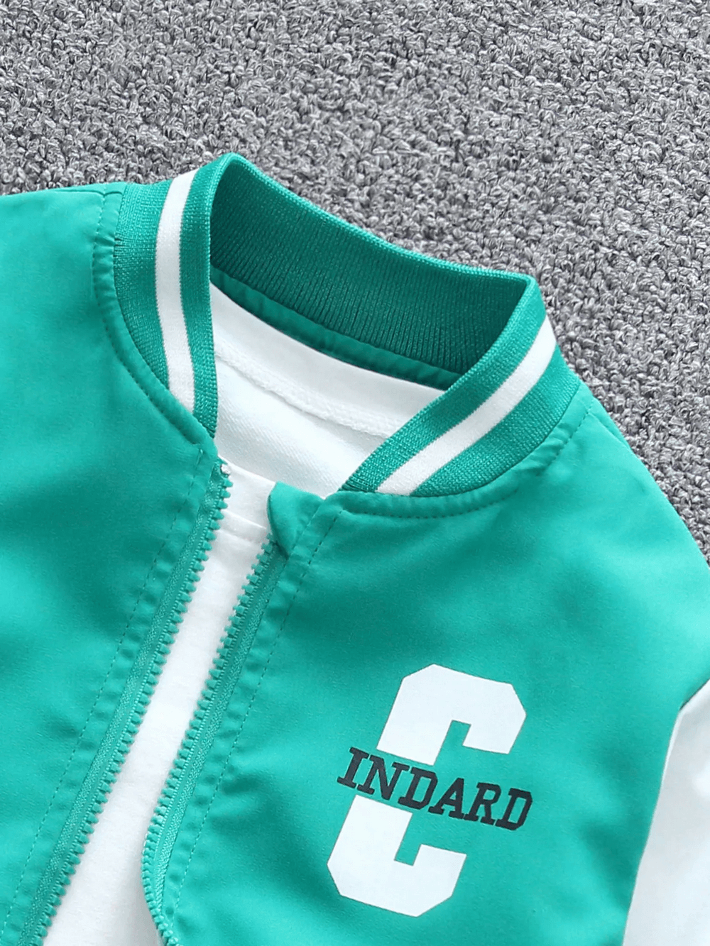 Kids varsity bomber jacket in teal with bold 'C' graphic, sporty design, white ribbed collar and zip-up front on grey background.