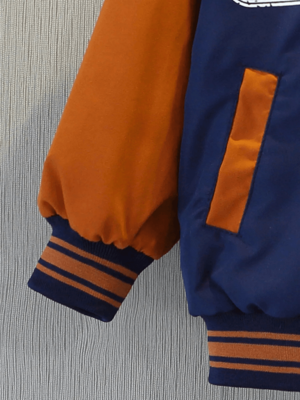 Close-up of kids varsity style jacket sleeve with orange and blue design, featuring sporty cuffs and pocket detailing.