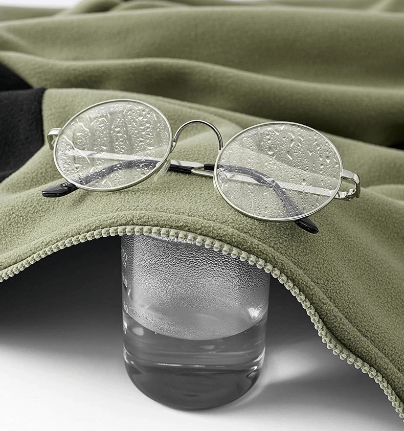 Round glasses on green fleece jacket with zipper and glass of water, showcasing warmth and casual style of unisex sports wear.