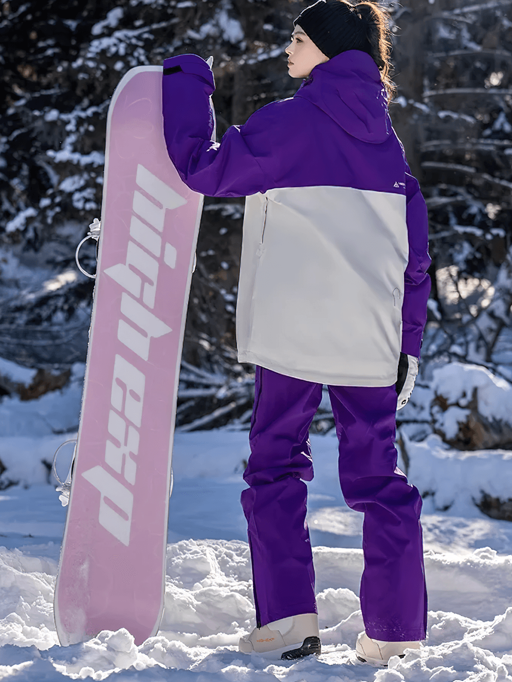 Woman in purple waterproof snowboarding jacket and pants set holding a snowboard in snowy mountains.