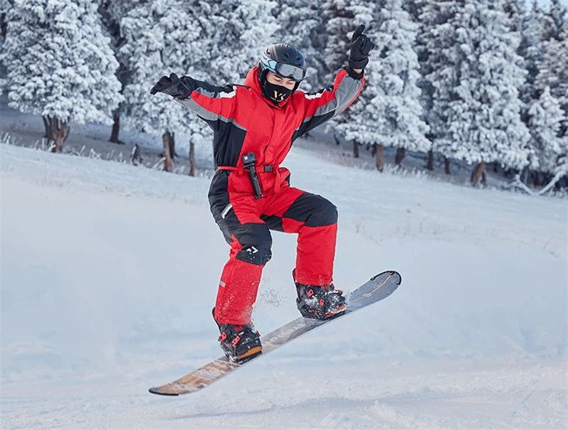 Snowboarder wearing a red waterproof warm ski jumpsuit mid-air in snowy forest, showcasing windproof snowboarding overalls for men.