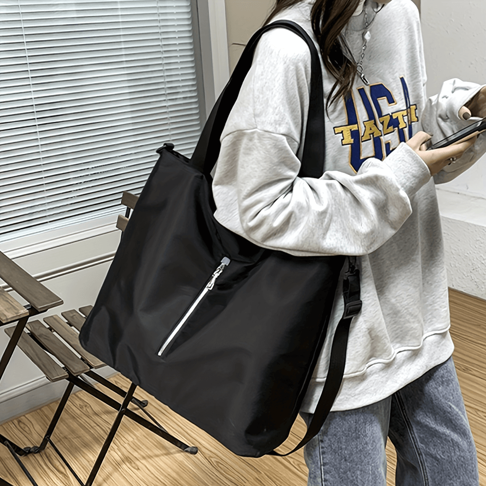 Woman wearing a grey sweatshirt with a large black nylon tote bag, featuring an adjustable strap and zip closure.